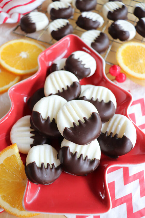 A red, tree-shaped dish holds several white shortbread cookies half dipped in chocolate. More cookies cool on a rack in the background, with orange slices and red berries nearby.