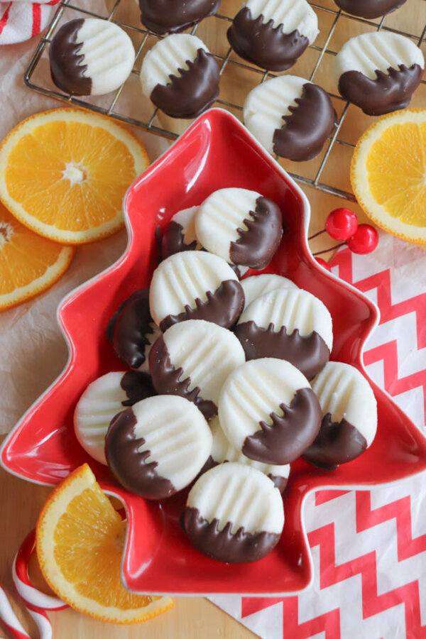 A red, tree-shaped dish holds round white cookies half-dipped in chocolate, surrounded by orange slices. More cookies rest on a cooling rack nearby. A red and white zigzag napkin is underneath.