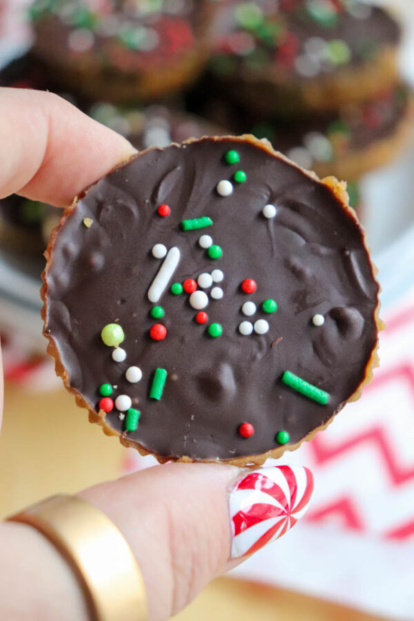 A hand with festive candy cane nail art holds a chocolate treat topped with colorful red, green, and white sprinkles. More treats are blurred in the background.