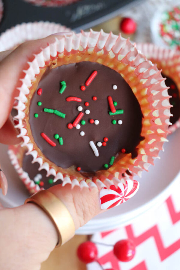 A hand holding a chocolate-covered treat in a muffin liner, topped with red, white, and green sprinkles. Festive decorations and other treats are visible in the background.