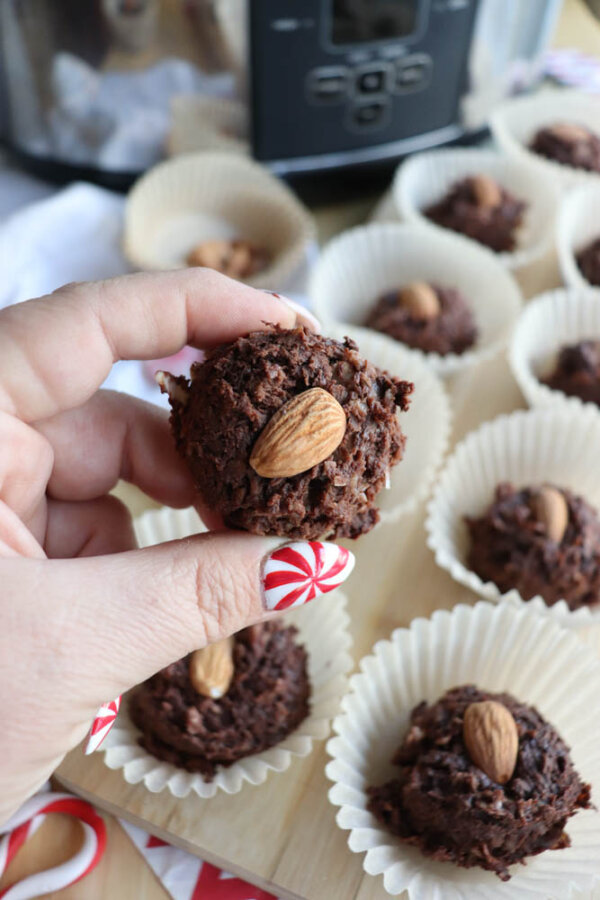 A hand holding a chocolate cookie topped with an almond above a tray of similar cookies in white paper liners. The person has red and white striped nail art. A kitchen appliance is visible in the background.
