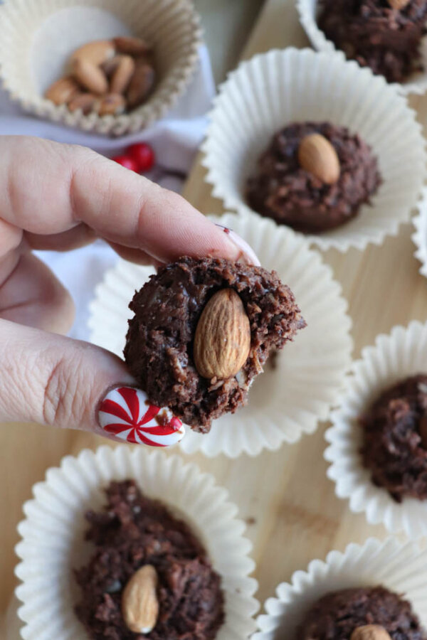 A hand with red and white peppermint nail art holds a chocolate almond treat with a bite taken out, showing the almond inside. Several more chocolate treats with almonds are arranged in white paper cups below.