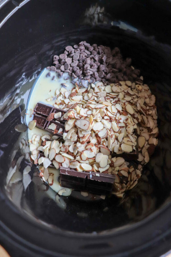 A close-up of a slow cooker filled with chocolate chips, chocolate squares, sliced almonds, and sweetened condensed milk, ready to be mixed and melted.