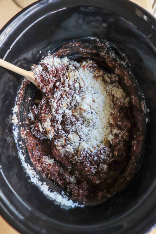 A black slow cooker filled with a chocolate mixture, shredded coconut, and chopped nuts being stirred with a wooden spoon.