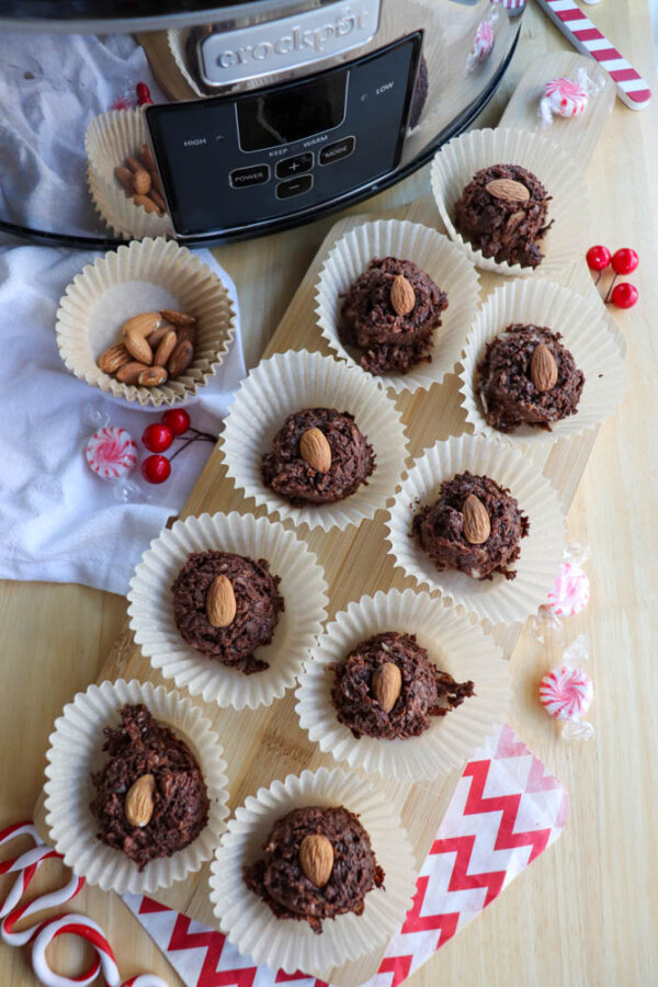 Chocolate almond clusters in paper liners are arranged on a wooden board near a slow cooker. There are whole almonds, candies, and festive napkins nearby, creating a holiday-themed dessert display.
