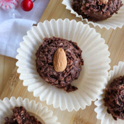 Chocolate cookies topped with whole almonds sit in white paper liners on a wooden tray. In the background, there are red cherries and a peppermint candy placed on a white cloth.