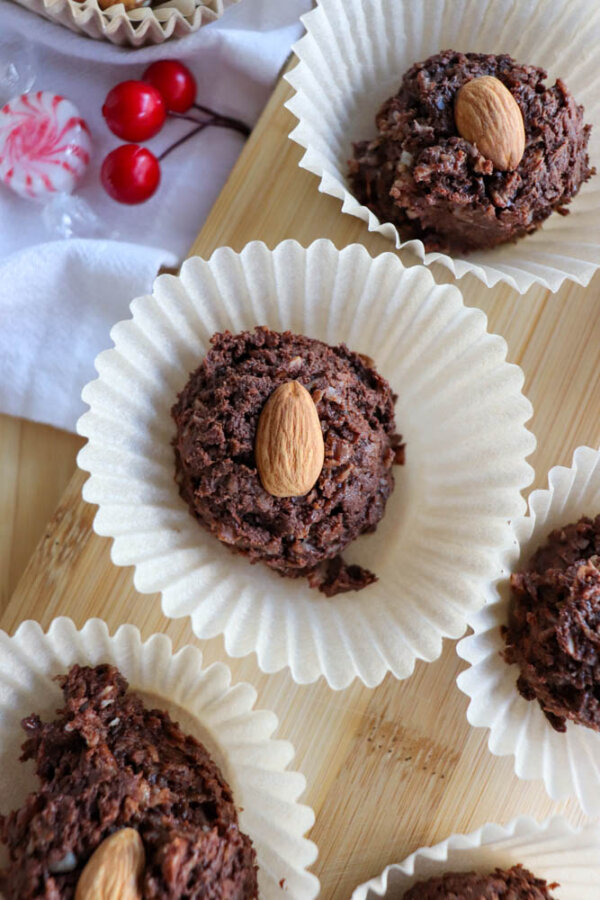 Chocolate cookies topped with whole almonds sit in white paper liners on a wooden tray. In the background, there are red cherries and a peppermint candy placed on a white cloth.