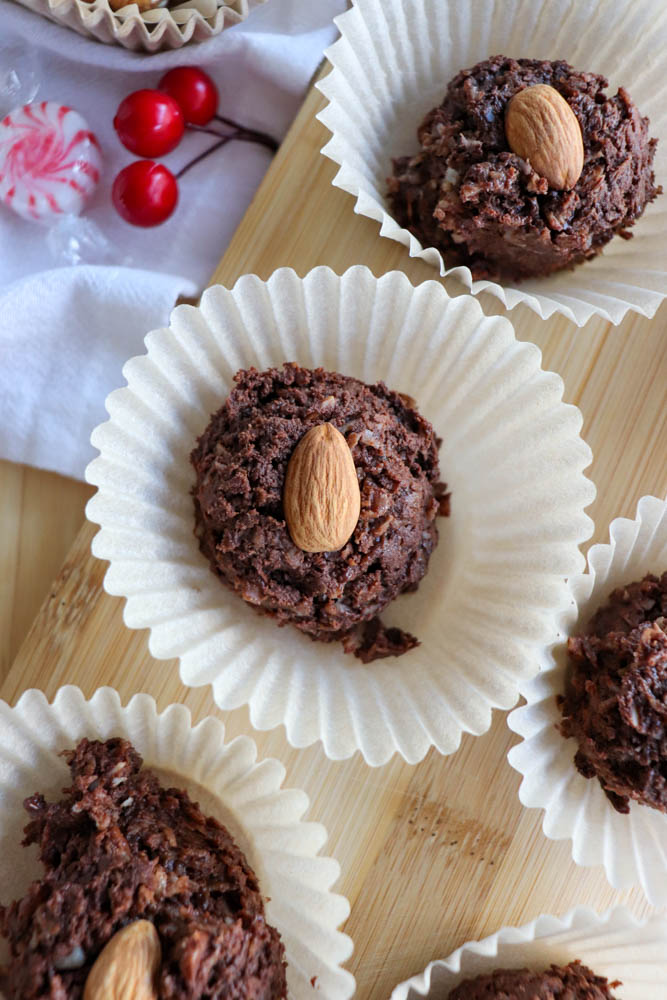 Chocolate cookies topped with whole almonds sit in white paper liners on a wooden tray. In the background, there are red cherries and a peppermint candy placed on a white cloth.