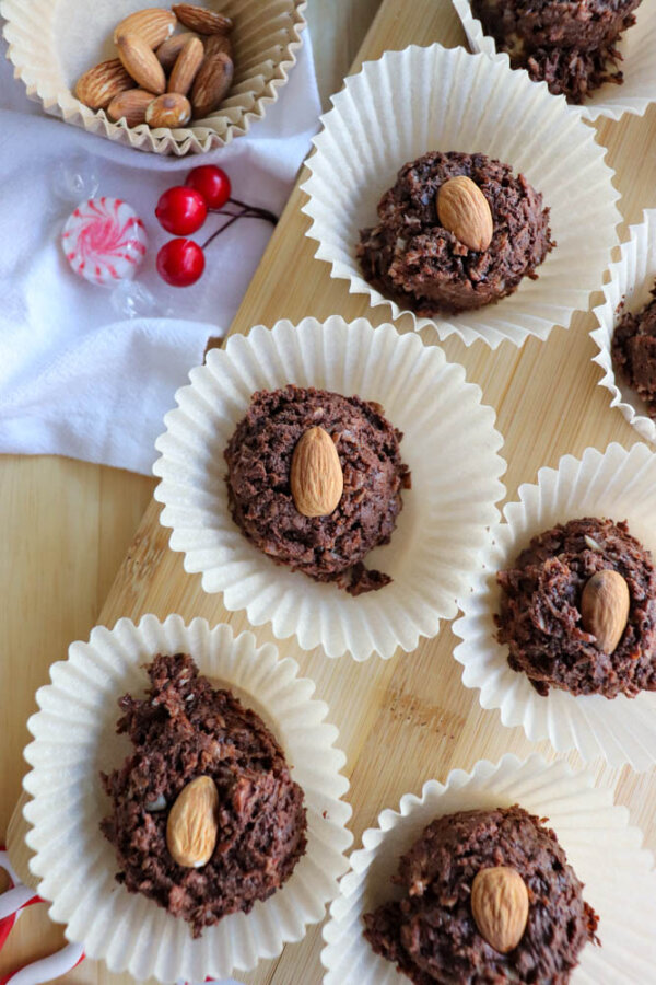 Chocolate almond cookies in paper liners arranged on a wooden surface, each topped with a whole almond. A bowl of almonds, peppermint candies, and red berries are nearby for decoration.
