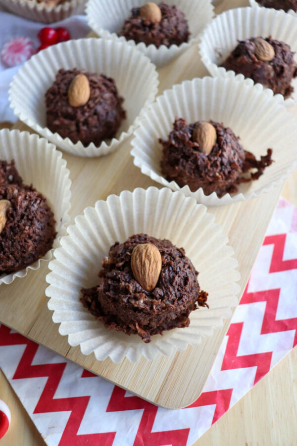 Chocolate coconut cookies in white paper liners, each topped with a whole almond, arranged on a wooden board over a red and white zigzag-patterned napkin.