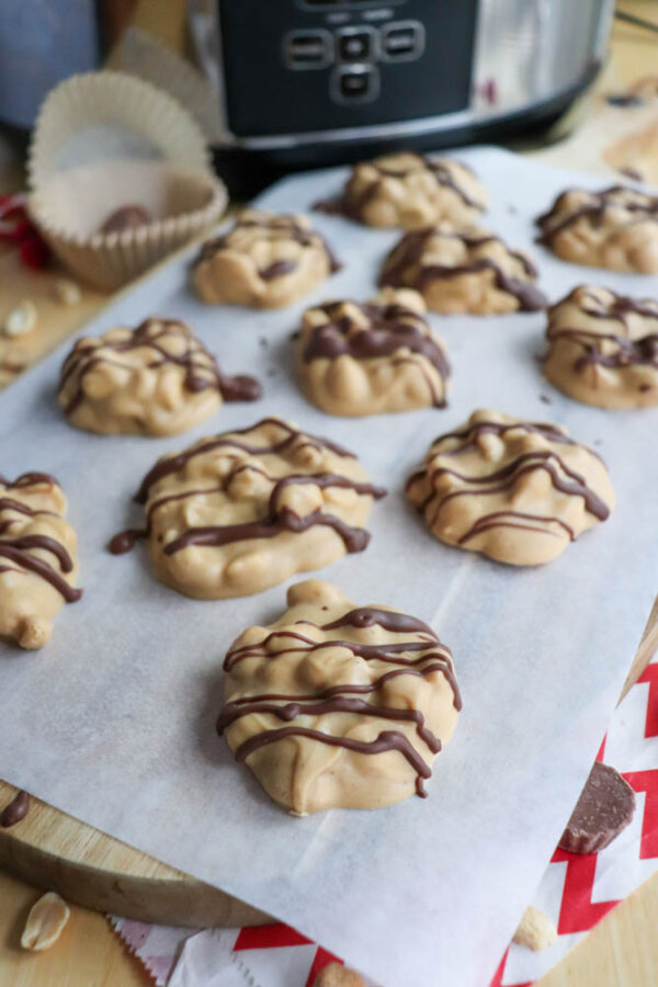 Cookies with a creamy, light brown coating and chocolate drizzle are arranged on parchment paper, with some candy wrappers, peanuts, and a slow cooker visible in the background.