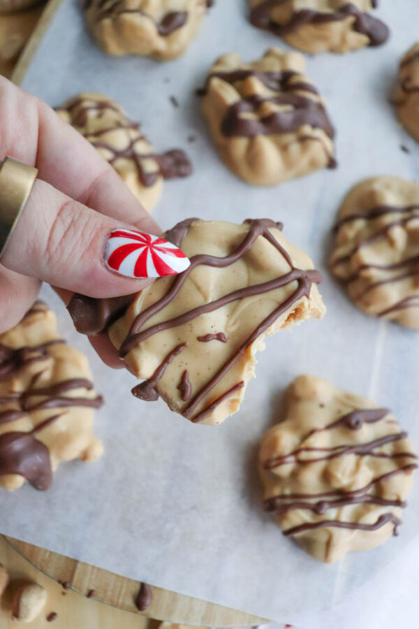 A hand with red and white striped nail art holds a cookie drizzled with chocolate, showing a bite taken out of it. More cookies are on parchment paper in the background.