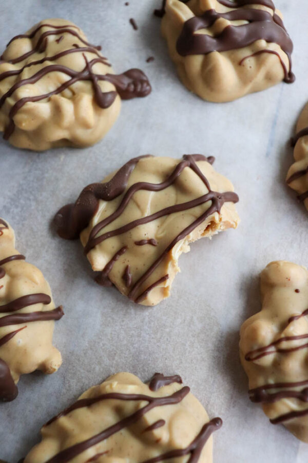 Close-up of several clusters of caramel and nuts coated in beige-colored candy and drizzled with dark chocolate, arranged on parchment paper. One candy piece has a bite taken out of it.