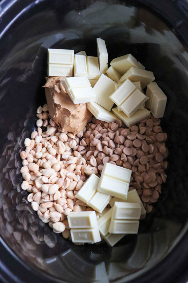 A close-up of a slow cooker containing white chocolate chunks, peanut butter, salted peanuts, and peanut butter chips before mixing.