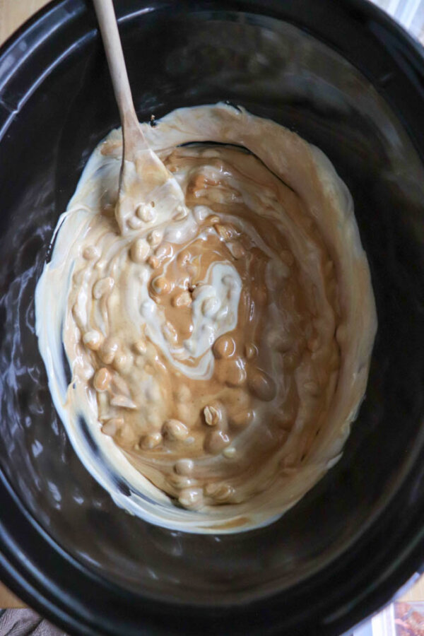 A wooden spoon stirs a creamy mixture of white and peanut butter chips in a black slow cooker. The ingredients are partially melted and swirled together.