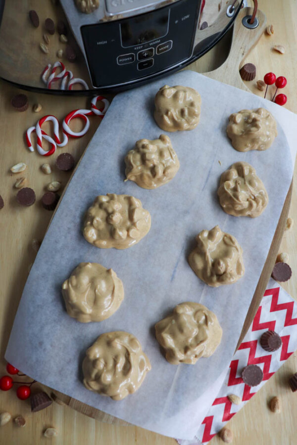 A sheet of parchment paper holds ten clusters of peanut candy. Surrounding the candy are peanuts, mini peanut butter cups, red and white peppermint candies, red candies, and a chevron-patterned napkin.