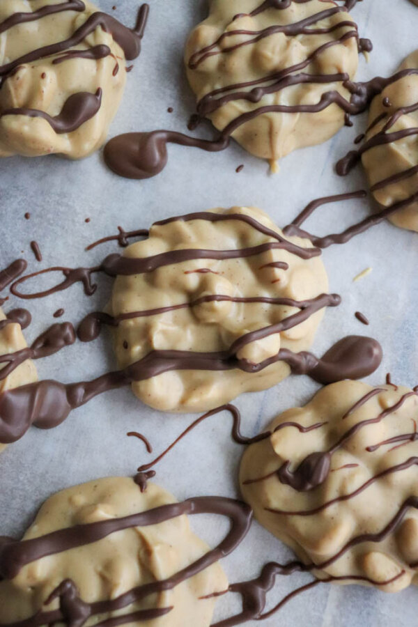 Five clusters of white candy coated treats with scattered peanuts are drizzled with chocolate and arranged on white parchment paper. The chocolate drizzle is uneven and artistic, covering parts of the clusters.