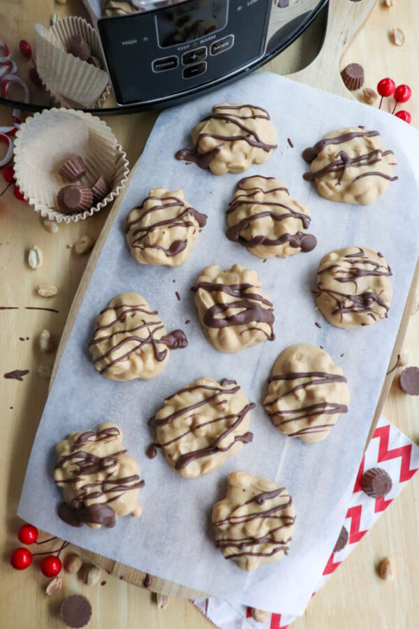 A tray of peanut butter clusters drizzled with chocolate sits on parchment paper. Surrounding the tray are cupcake liners, peanuts, mini peanut butter cups, and red candy-coated chocolates on a wooden surface.