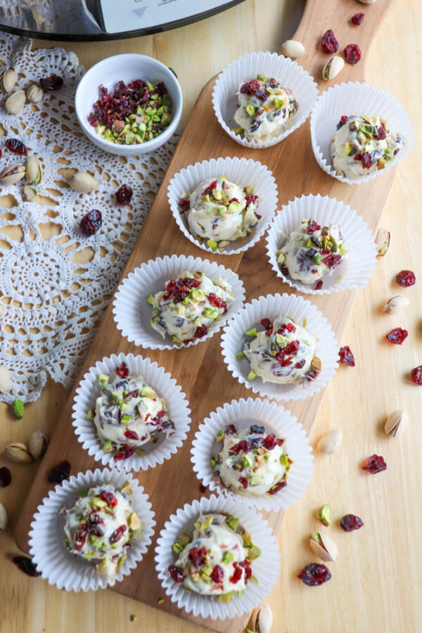 Ten white paper cupcake liners filled with clusters of white chocolate, dried cranberries, and chopped pistachios are arranged on a wooden board, with extra toppings scattered around and a doily partially visible.