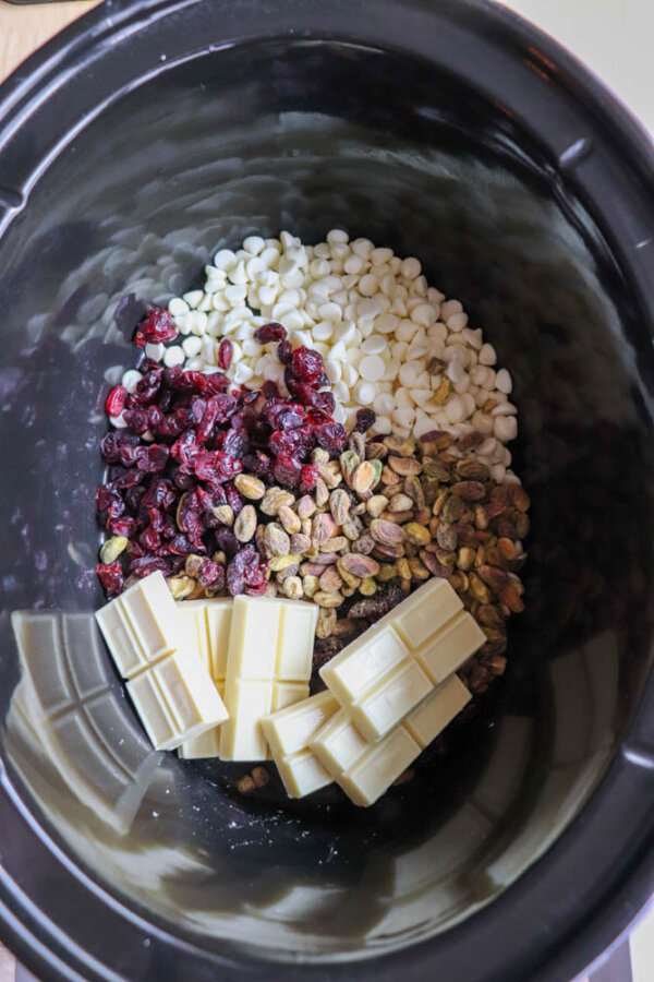A slow cooker filled with white chocolate chips, dried cranberries, pistachios, and white chocolate bars, each ingredient separated in its own section.