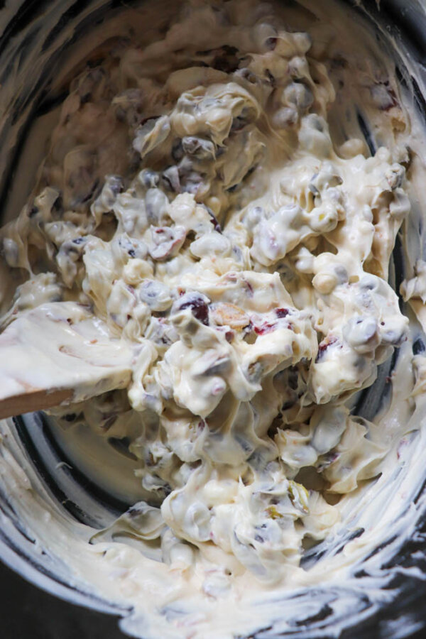 A close-up of a creamy mixture with white coating, nuts, and dried fruit being stirred with a spatula in a bowl.