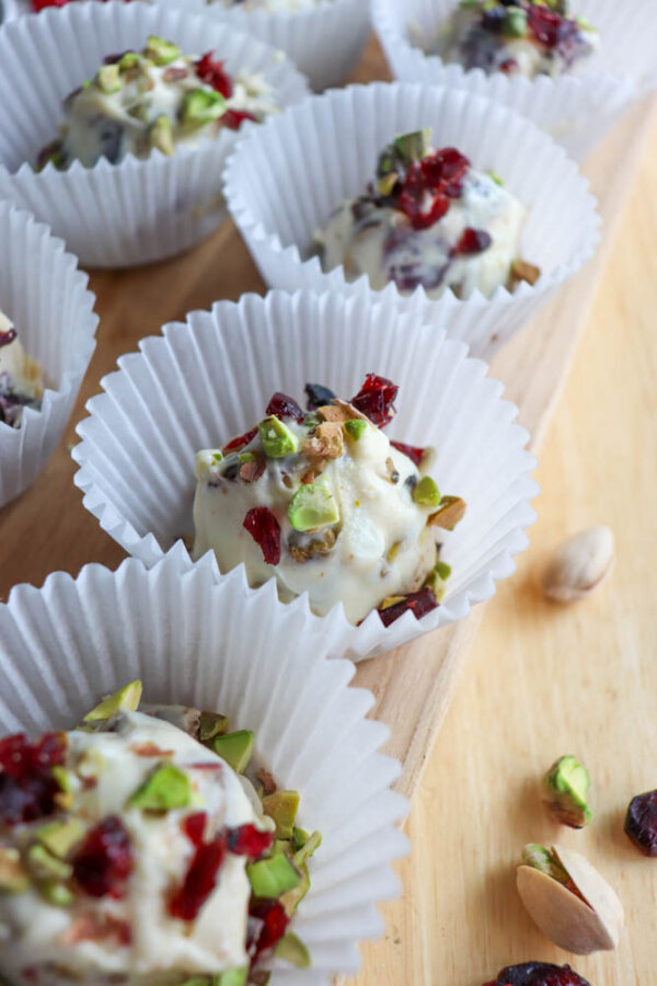 White chocolate clusters topped with chopped pistachios and dried cranberries sit in white paper cupcake liners on a wooden surface. A few pieces of pistachio and cranberry are scattered nearby.
