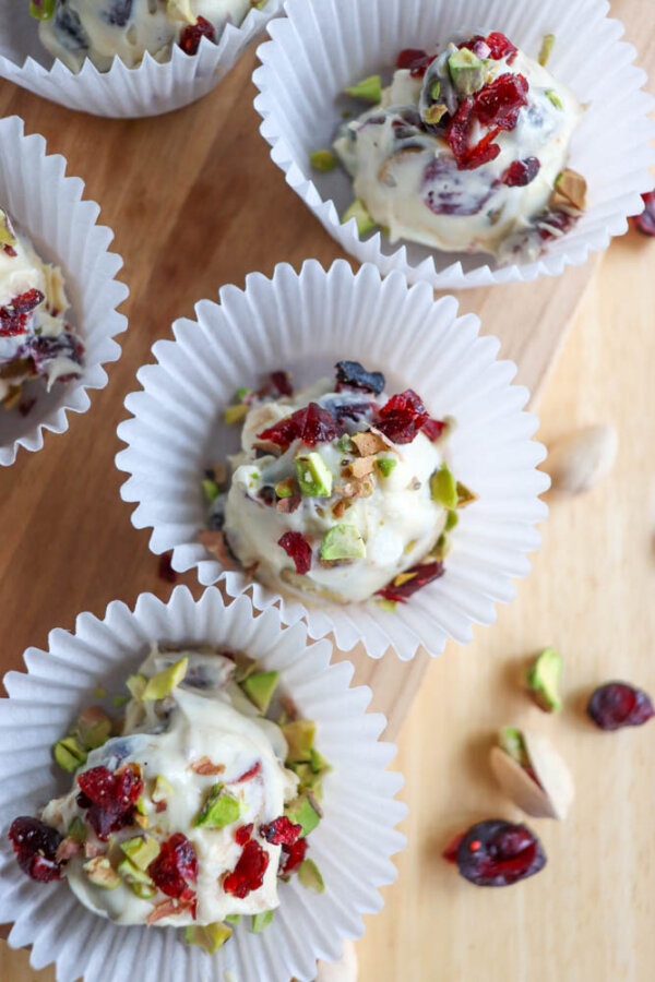 Small clusters of white chocolate with chopped pistachios and dried cranberries, placed in white paper cups on a wooden surface. Some pieces of pistachio and cranberry are scattered nearby.