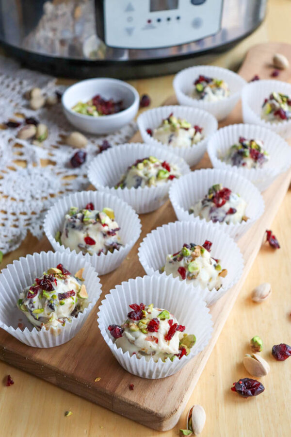 A wooden board displays white paper cups filled with white chocolate truffles topped with chopped pistachios and dried cranberries. More nuts and cranberries are scattered around, with a slow cooker in the background.