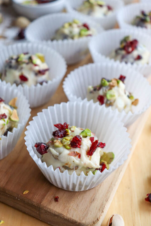 Several white paper cupcake liners hold clusters of white chocolate coated nuts, topped with chopped pistachios and dried cranberries, arranged on a wooden surface.