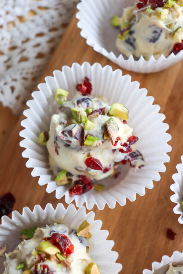 A close-up of white chocolate truffles in paper liners, topped with chopped pistachios and dried cranberries, arranged on a wooden surface.