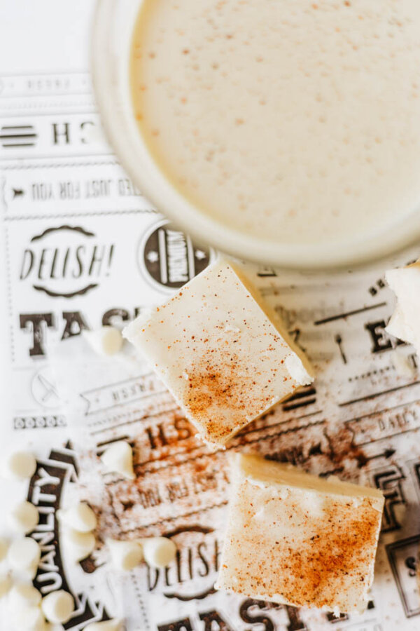Three creamy fudge squares sprinkled with cinnamon sit next to scattered white chocolate chips and a glass of light-colored drink, all placed on a decorative black and white paper.