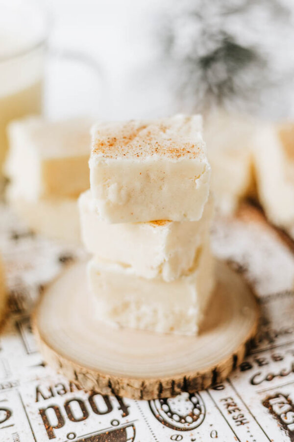 Three creamy white fudge squares are stacked on a small wooden slice, with a dusting of spice on top. More fudge pieces and a blurred background are visible behind the stack.