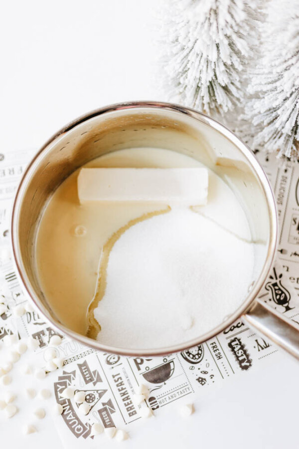 A saucepan filled with a stick of butter, granulated sugar, and sweetened condensed milk, placed on a decorative paper with snow-tipped artificial trees nearby.