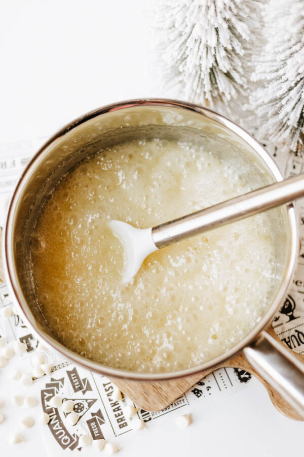 A saucepan filled with a bubbling, light-colored mixture being stirred with a white spatula, set on a wooden board next to a snowy artificial pine branch and a sheet with small white chocolate chips.