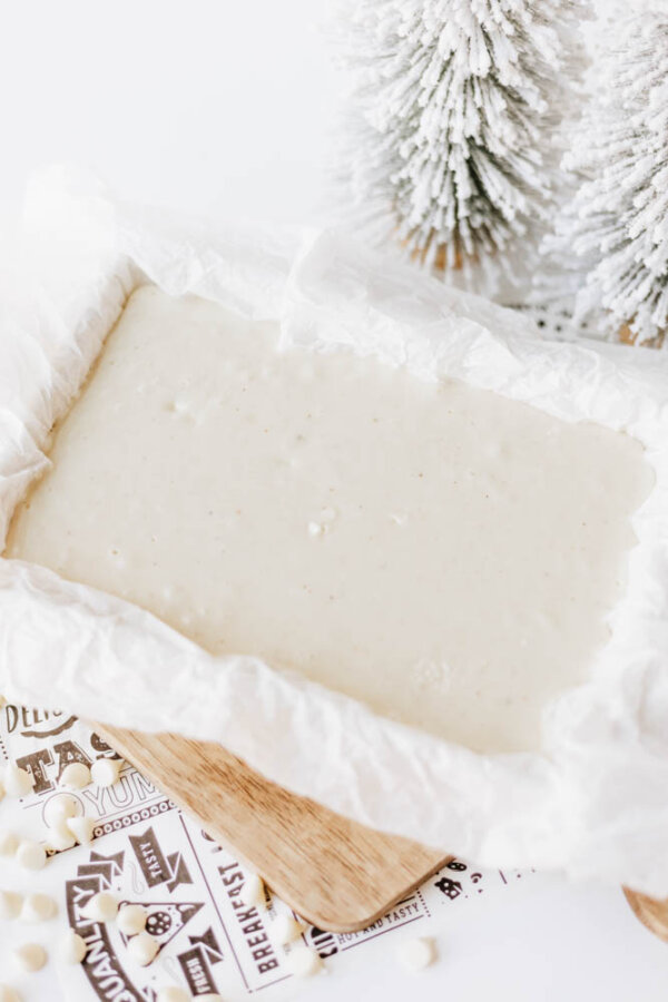A baking dish lined with parchment paper is filled with white chocolate fudge mixture, set on a wooden board with two snowy decorative trees and white chocolate chips nearby.