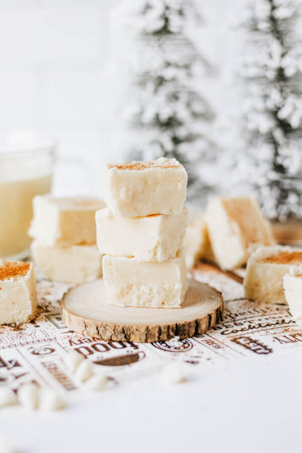 Three pieces of white fudge are stacked on a round wooden slice, surrounded by more fudge pieces. The background features blurred snowy pine trees and a light festive setting.
