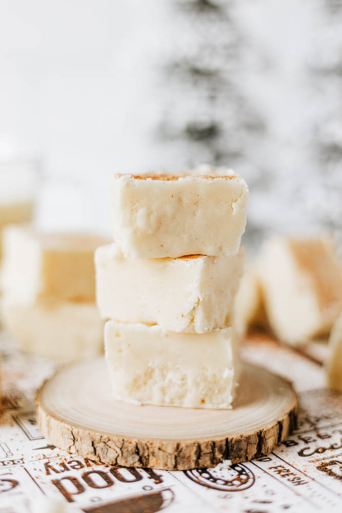 Three creamy white fudge squares are stacked on a round wooden slice, with more fudge pieces blurred in the background. The setup rests on patterned paper with a rustic, festive feel.