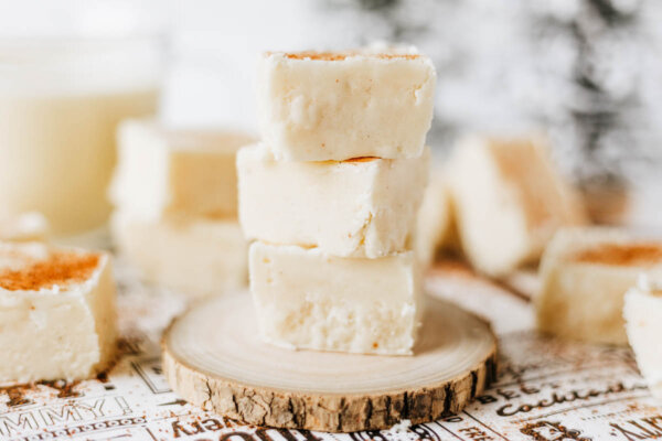 Squares of creamy white fudge, some stacked on a small wooden slab, with a light dusting of cinnamon on top, set against a blurred background and festive surface.