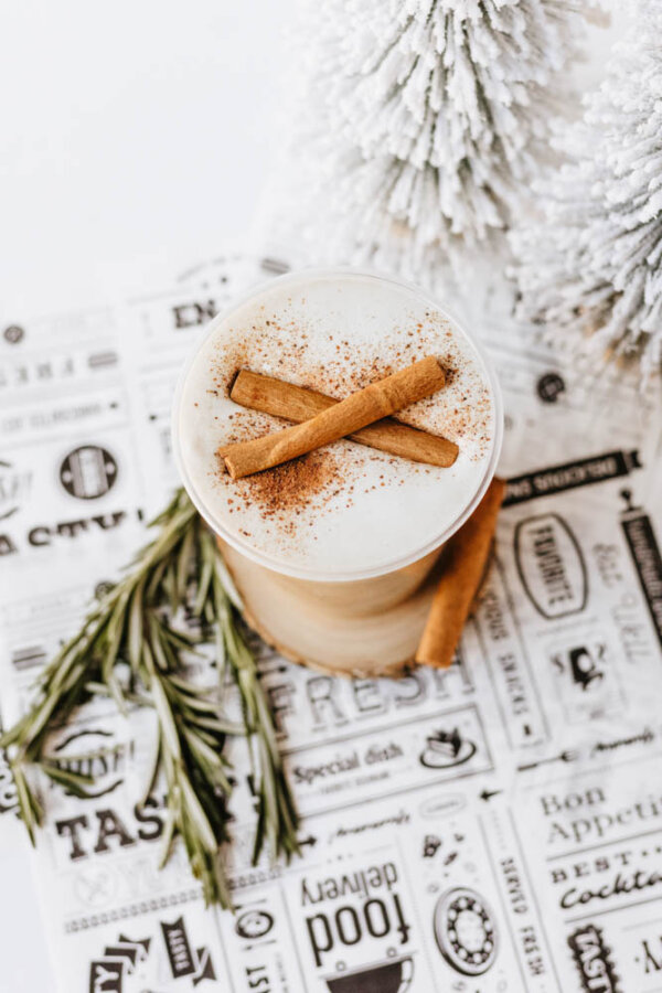 A festive drink topped with frothy cream, two cinnamon sticks, and a sprinkle of cinnamon, sits beside rosemary sprigs and snowy decorative trees on a white patterned surface.