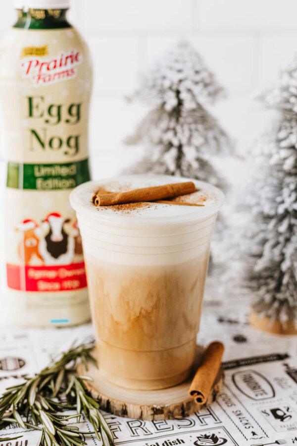 A plastic cup of iced eggnog latte topped with cinnamon sticks sits on a coaster, with a bottle of Prairie Farms Egg Nog and decorative snow-covered trees in the background.