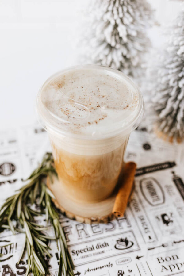 A plastic cup of iced coffee with foam and sprinkled spices on top sits on a decorative paper, with a sprig of rosemary and a cinnamon stick beside it. Snowy artificial trees are in the background.