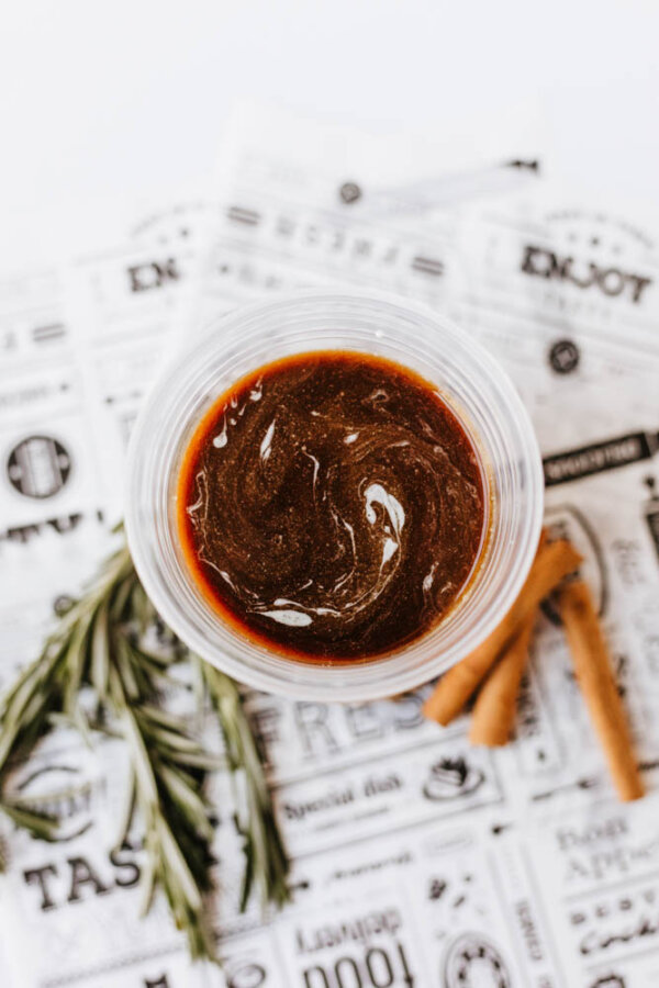 A clear cup filled with dark brown sauce sits on a sheet of printed paper, next to a sprig of rosemary and two cinnamon sticks.