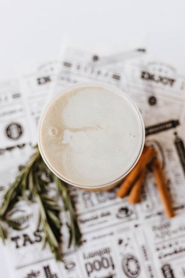A top-down view of a creamy latte in a clear glass, placed on decorative newspaper-like paper, with cinnamon sticks and rosemary sprigs nearby.