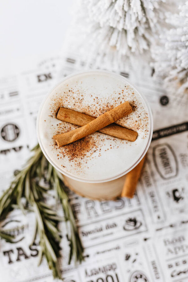 A creamy beverage topped with frothy foam, sprinkled with cinnamon and garnished with two cinnamon sticks, sits on a decorative paper next to a sprig of rosemary and a white festive decoration.