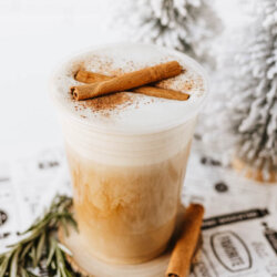 A creamy iced latte topped with foam and cinnamon sticks, garnished with rosemary, sits on a coaster next to decorative snowy trees. The scene has a cozy, wintery feel.
