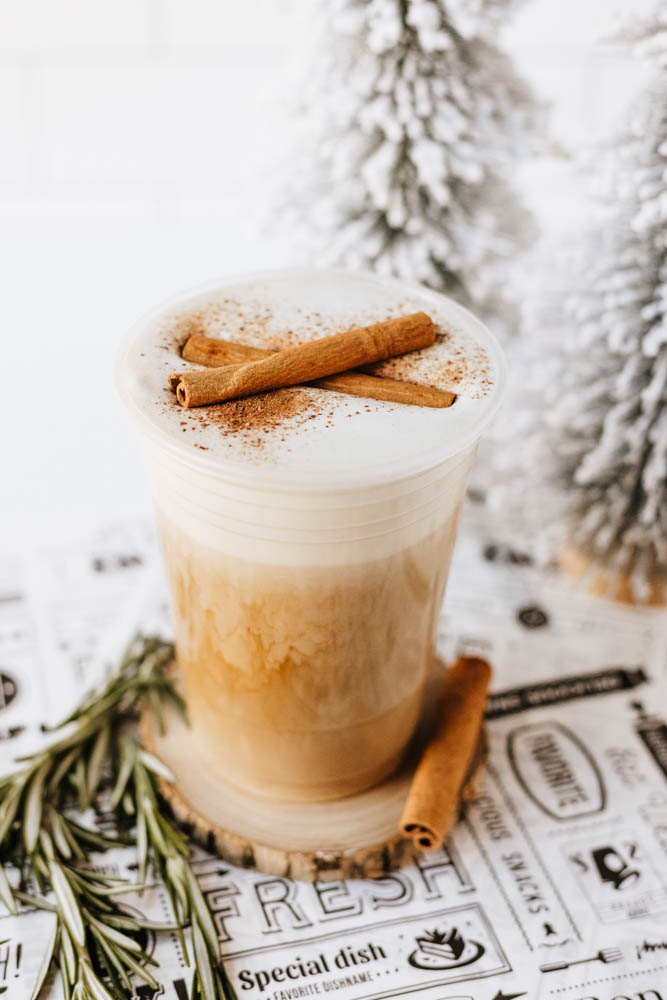 A creamy iced latte topped with foam and cinnamon sticks, garnished with rosemary, sits on a coaster next to decorative snowy trees. The scene has a cozy, wintery feel.