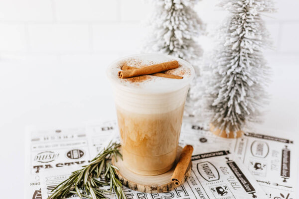 A cup of frothy coffee topped with cinnamon sticks sits next to a sprig of rosemary and cinnamon sticks, with two small, snow-covered decorative trees in the background.