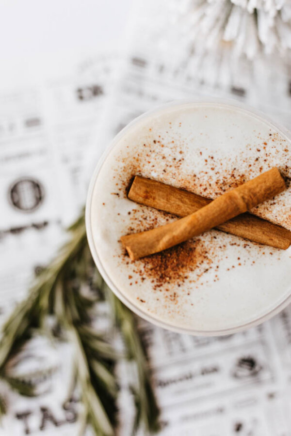 A creamy beverage topped with two cinnamon sticks and a sprinkle of ground cinnamon, set on a newspaper-patterned surface with a sprig of rosemary nearby.