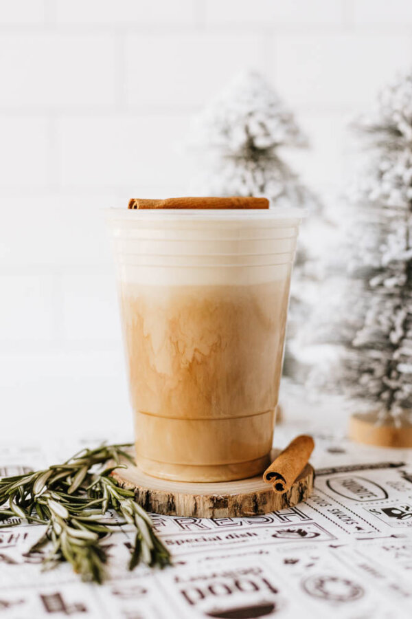 A plastic cup of iced latte with layered milk and coffee, topped with a cinnamon stick, sits on a wood slice coaster. There’s a sprig of rosemary and another cinnamon stick nearby, with frosted mini pine trees in the background.