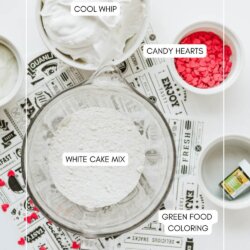 A flat lay of ingredients for a Grinch cookie dip recipe on a white surface, including a bowl of Cool Whip, white cake mix, small bowls of candy hearts, green food coloring, and a printed placemat underneath.
